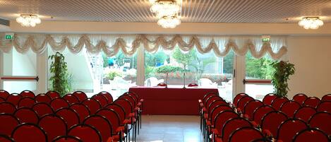 Une salle de conférence avec des rangées de chaises rouges, une table rouge et un plafond orné d’un motif en réseau et de lustres.