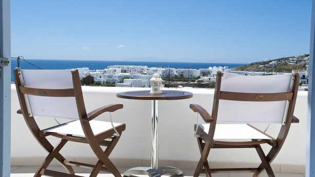 A balcony with two wooden chairs and a small round table, overlooking a coastal town with white buildings and a clear blue sky. - Villa Nireas (Mykonos)