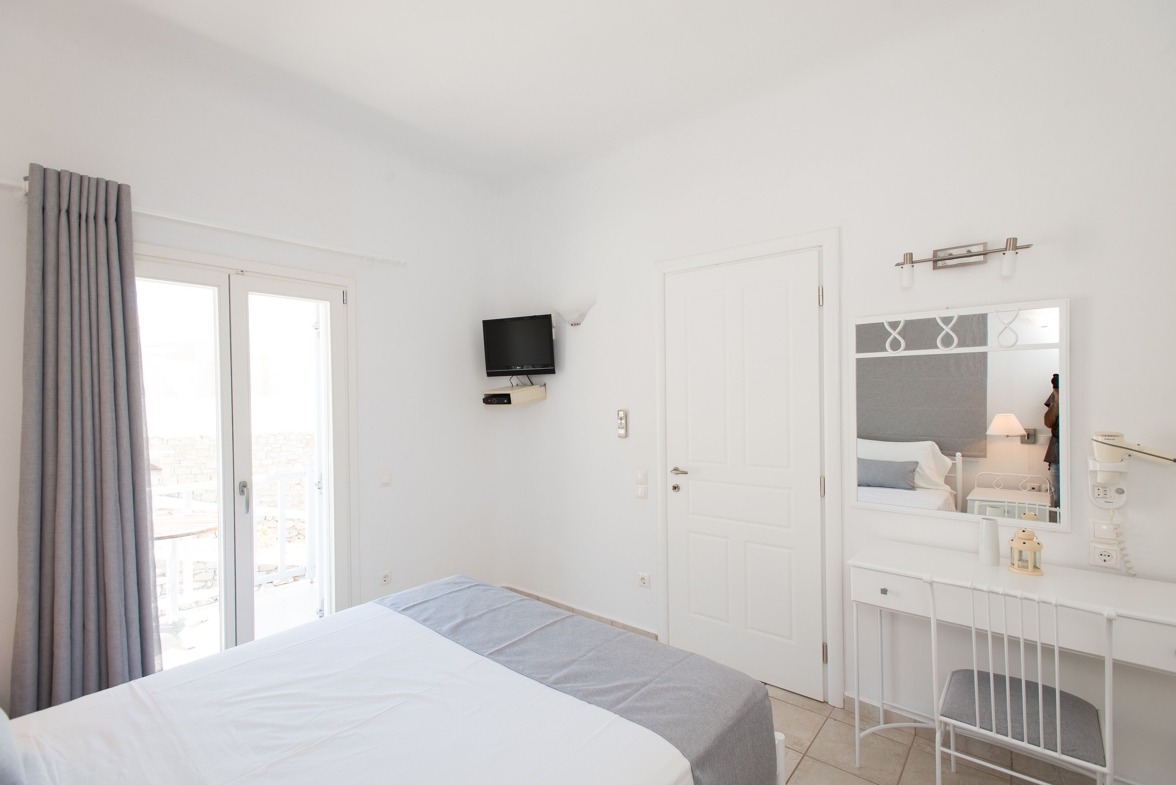 Een slaapkamer met een bed, een televisie aan de muur, een bureau met een spiegel, en een deur naar een buitenruimte.