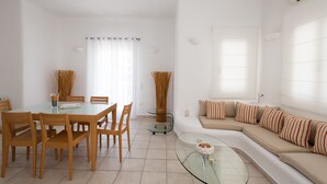 A modern living room with a glass dining table, wooden chairs, a glass coffee table, a beige sofa with striped cushions, and a large window with sheer curtains. - Villa Nireas (Mykonos)