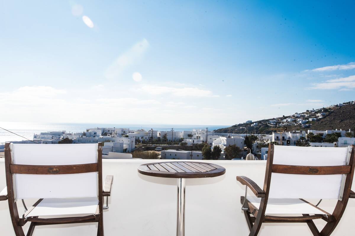 Two wooden chairs with white cushions, a small round table, and a view of a coastal town with white buildings and a clear sky.