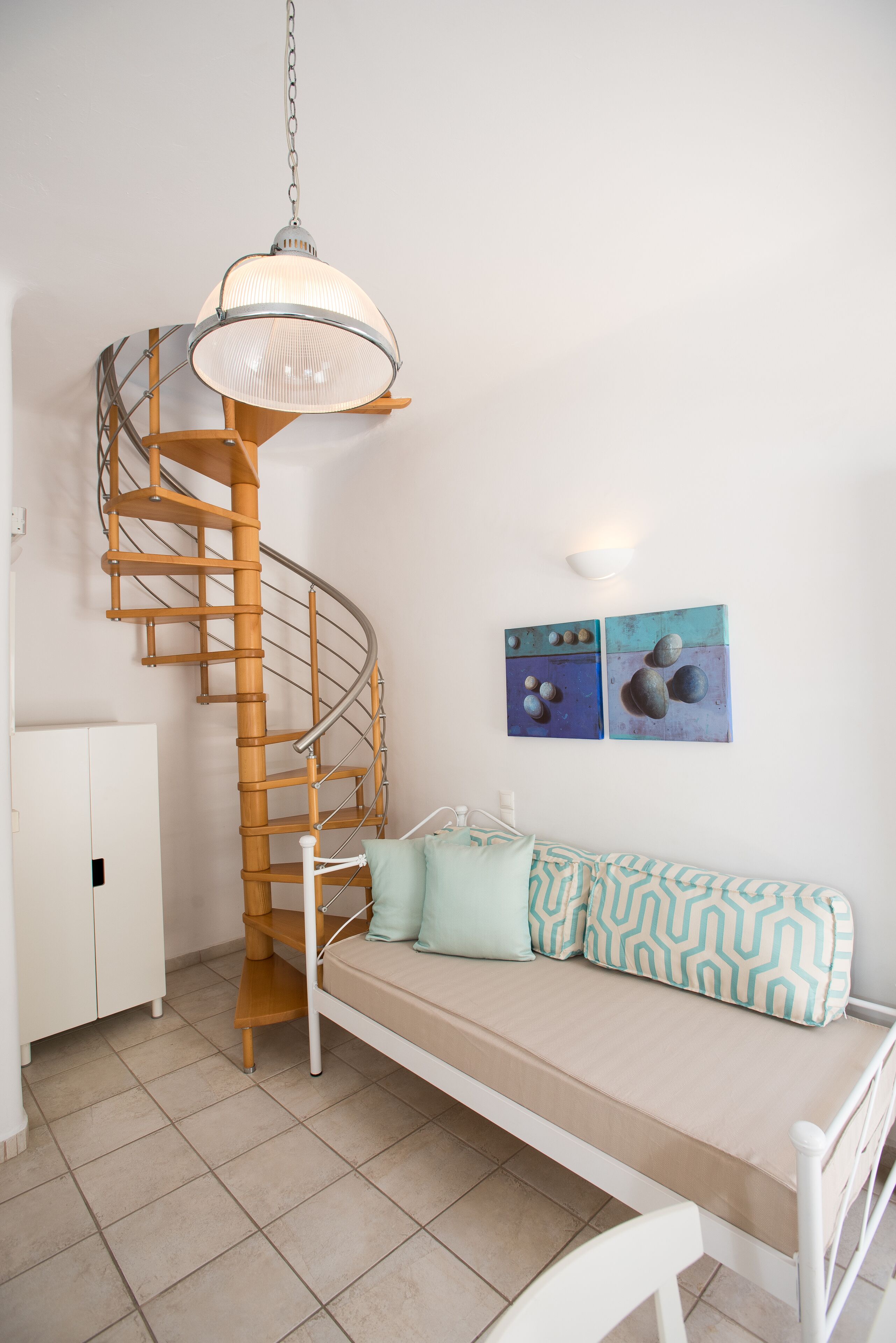 A spiral staircase with a white wall and a ceiling light. There is a white sofa with patterned cushions and a white chair. Two paintings are on the wall.
