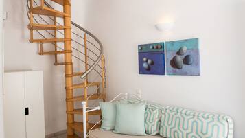 Una escalera de caracol con pared blanca y una lámpara en el techo. Hay un sofá blanco con cojines estampados y una silla blanca. Dos cuadros decoran la pared.