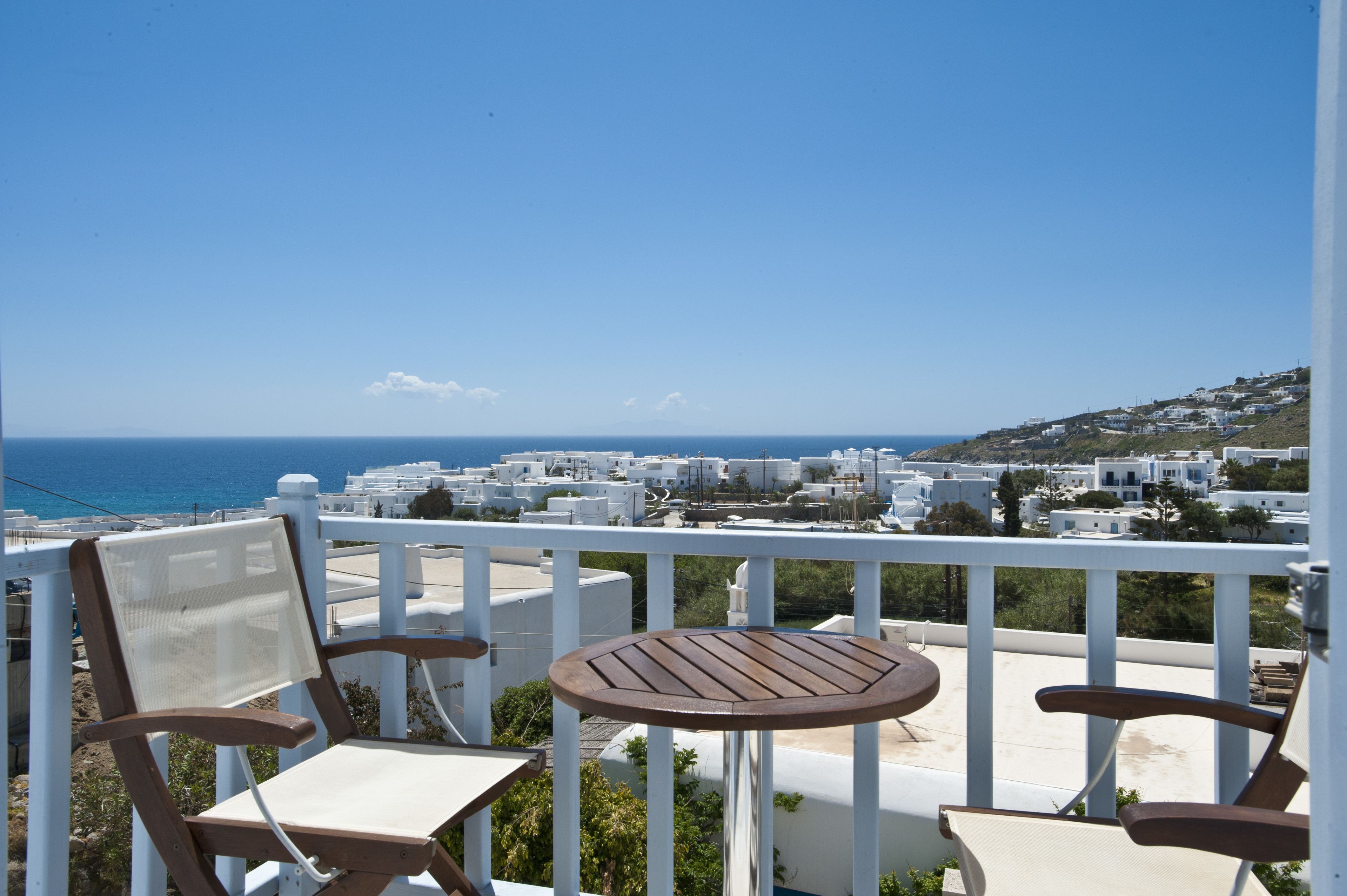 Un balcone con due sedie di legno e un tavolino, con vista su una cittadina costiera caratterizzata da edifici bianchi e dal mare.