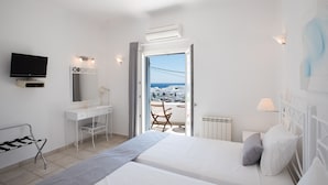 A modern hotel room with a bed, a desk, a chair, a television, and a balcony with a view of the city. - Villa Nireas (Mykonos)