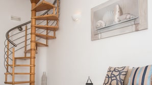 A modern living room with a wooden spiral staircase, a white sofa with patterned cushions, and a decorative wall piece. - Villa Nireas (Mykonos)
