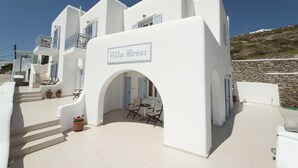 A white villa with a sign reading "Villa Nireas." It features a patio with a table and chairs, potted plants, and a stone wall. - Villa Nireas (Mykonos)