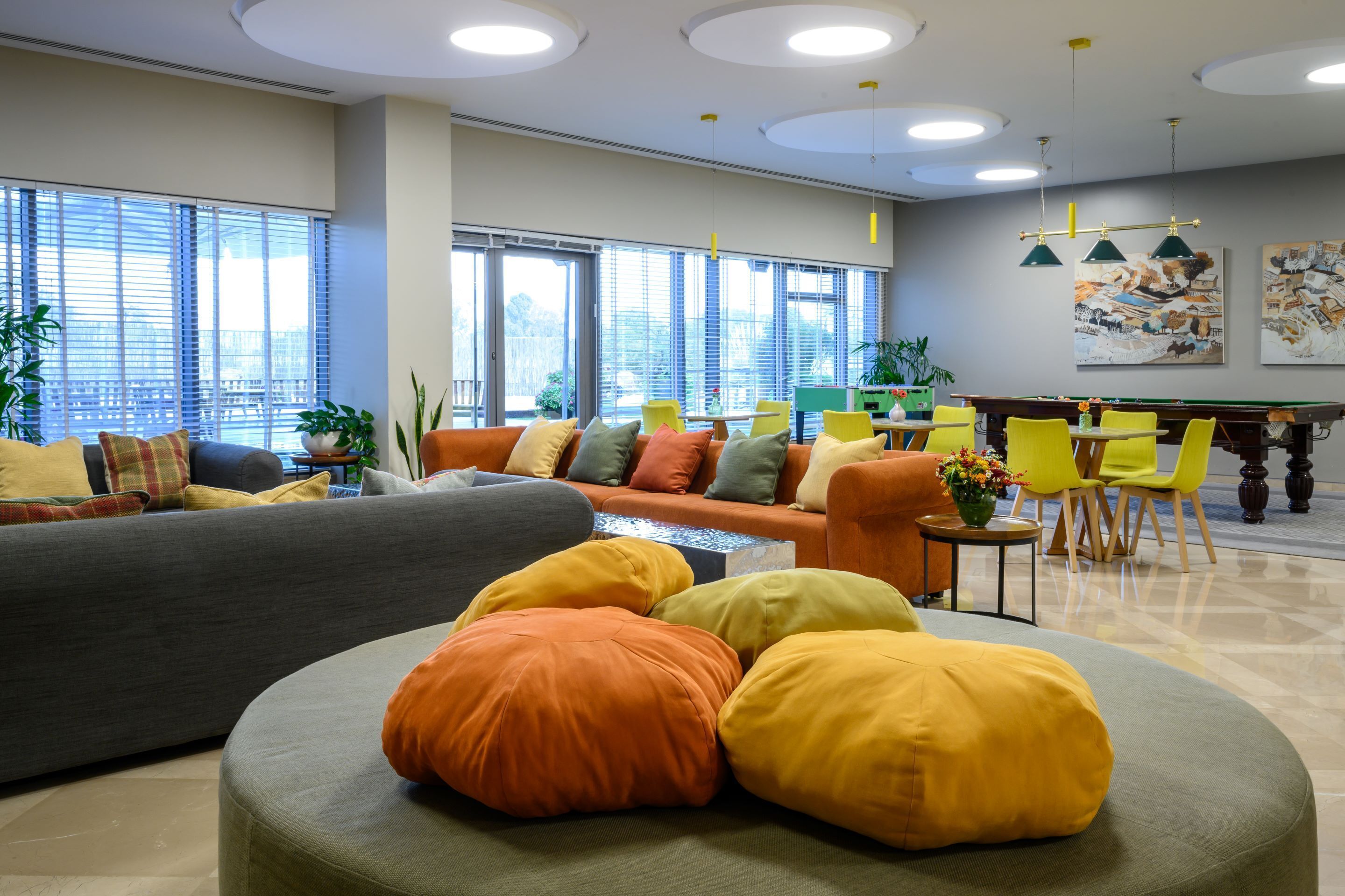 A modern lounge area with a sofa, ottoman, and chairs, a pool table, and large windows with blinds.