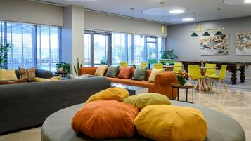 Un área de descanso moderna con sofá, reposapiés y sillones, mesa de billar y ventanas amplias con persianas.