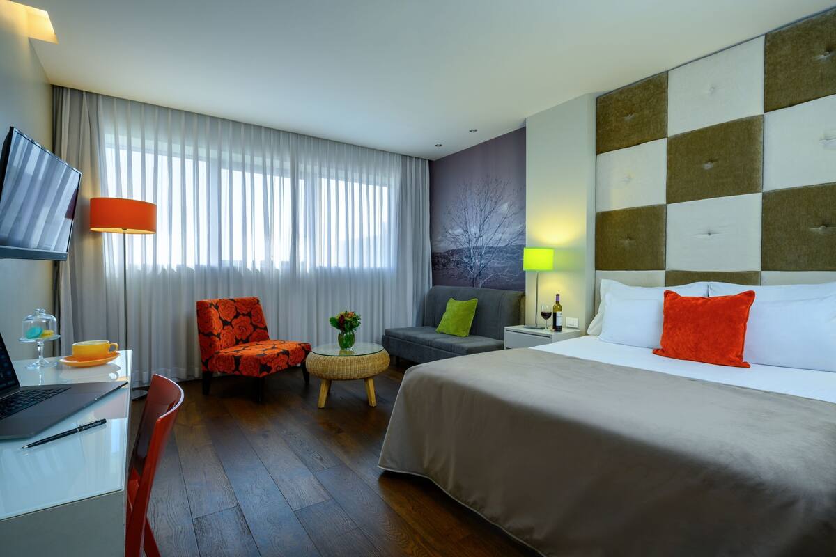 A hotel room with a bed, a desk with a laptop, a chair, a small table with a vase, and a window with curtains.