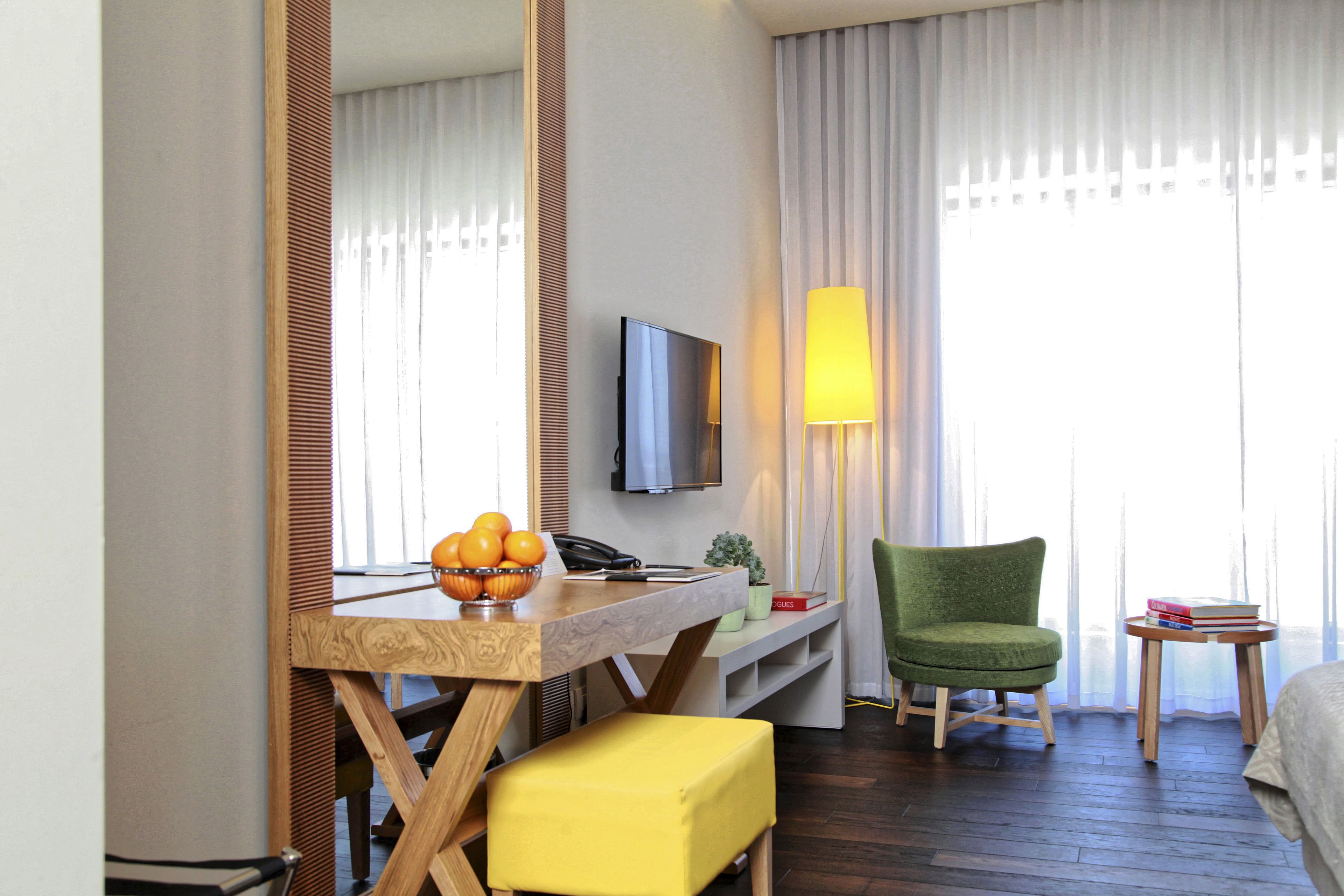 Une chambre d’hôtel avec un bureau en bois, une télévision, un fauteuil vert, un pouf jaune et une petite table avec des livres.
