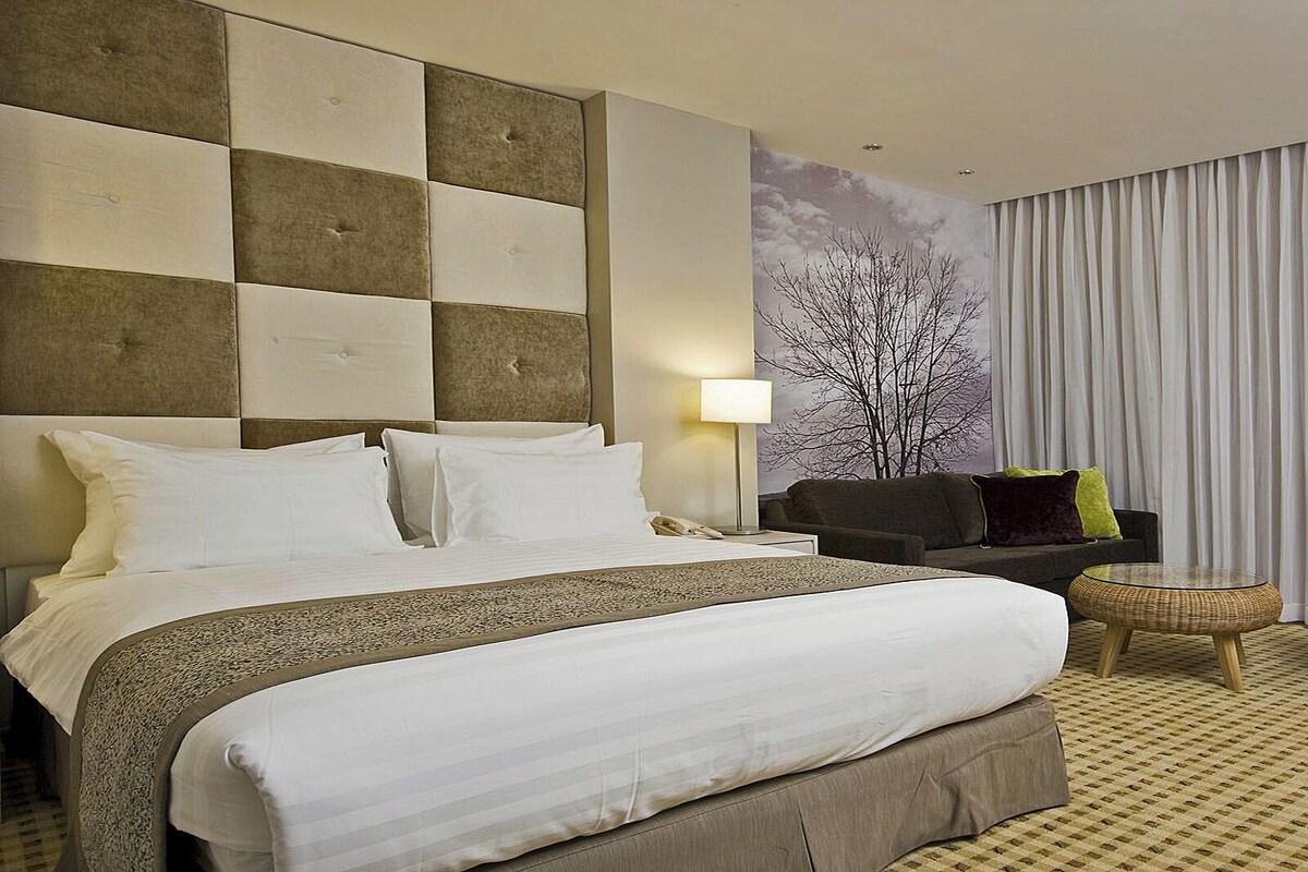 A hotel room with a large bed, a sofa, a chair, and a wall with a tree mural.