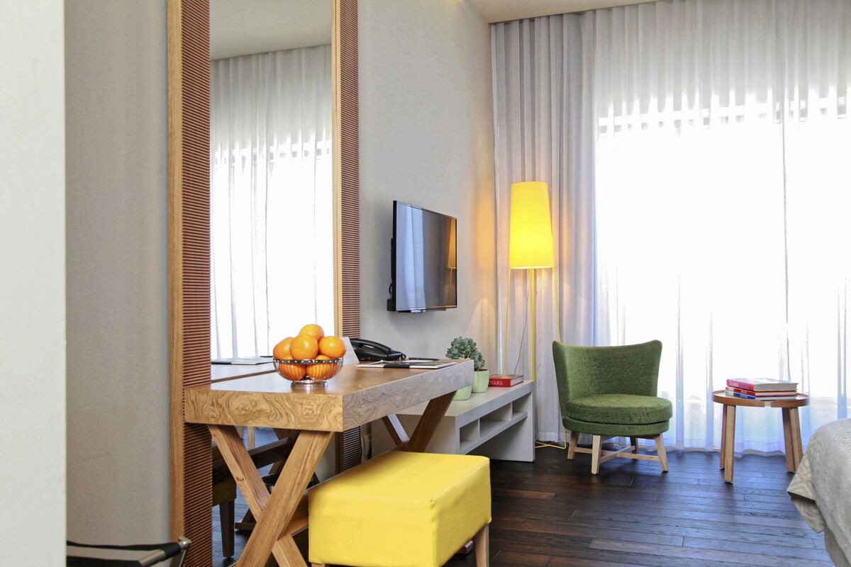 A hotel room with a wooden desk, a green chair, a yellow ottoman, a television, and a lamp.