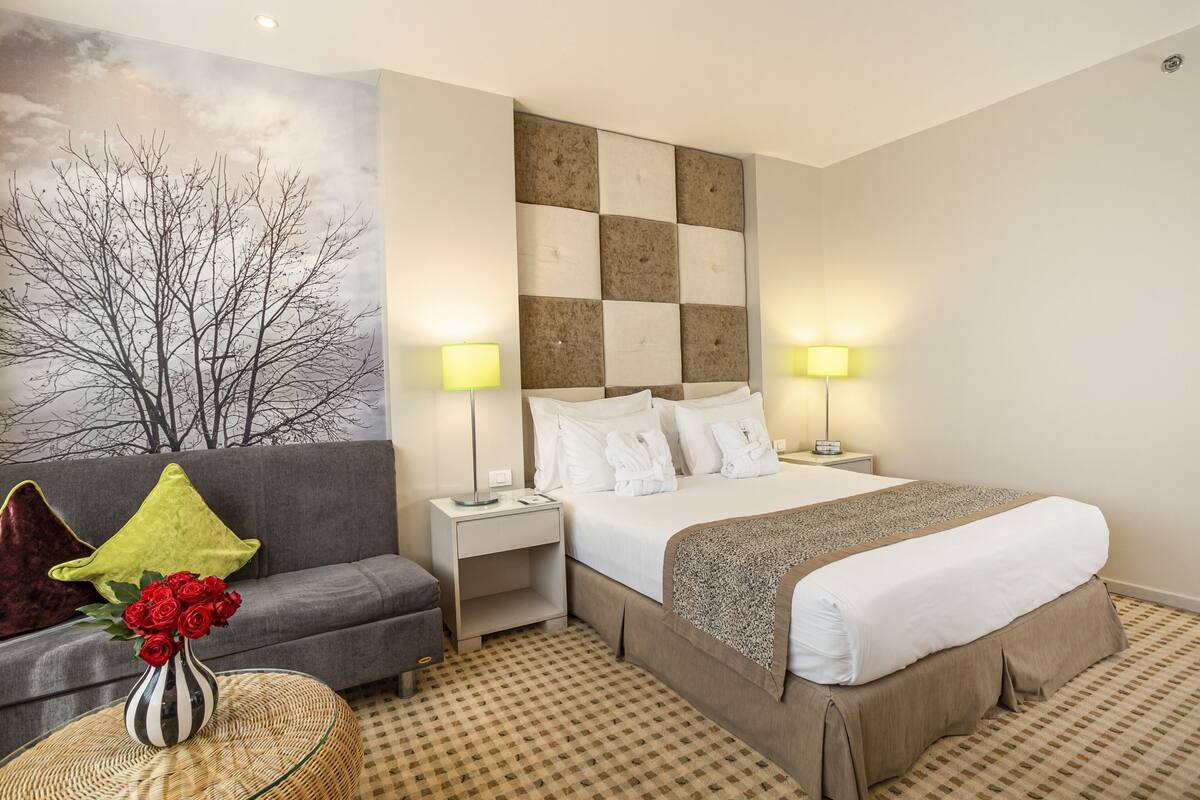 A hotel room with a bed, a sofa, a nightstand, a lamp, and a wall with a tree mural.