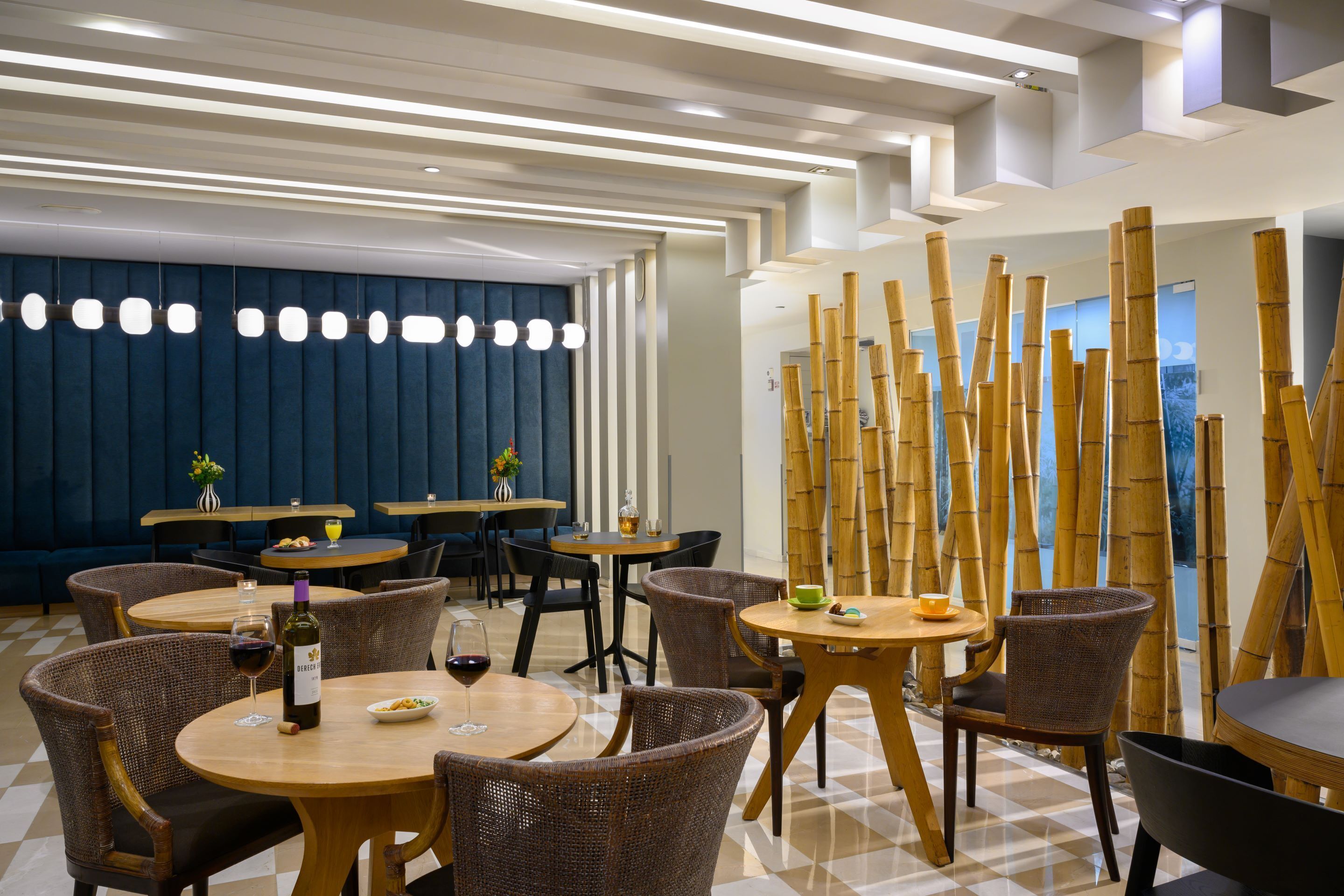 Un restaurante moderno con mesas redondas y sillas de mimbre, con piso de cuadros y pared azul con líneas verticales.