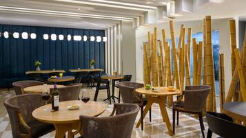 Un restaurante moderno con mesas redondas y sillas de mimbre, con piso de cuadros y pared azul con líneas verticales.
