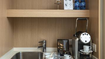 Une cuisine compacte équipée d’un évier en acier inoxydable, d’une machine à café Nespresso et d’une étagère où sont rangées des bouteilles d’eau et un verre.