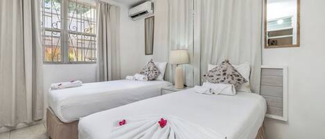 A hotel room with two beds, a ceiling fan, an air conditioning unit, a bedside lamp, and a window with curtains.