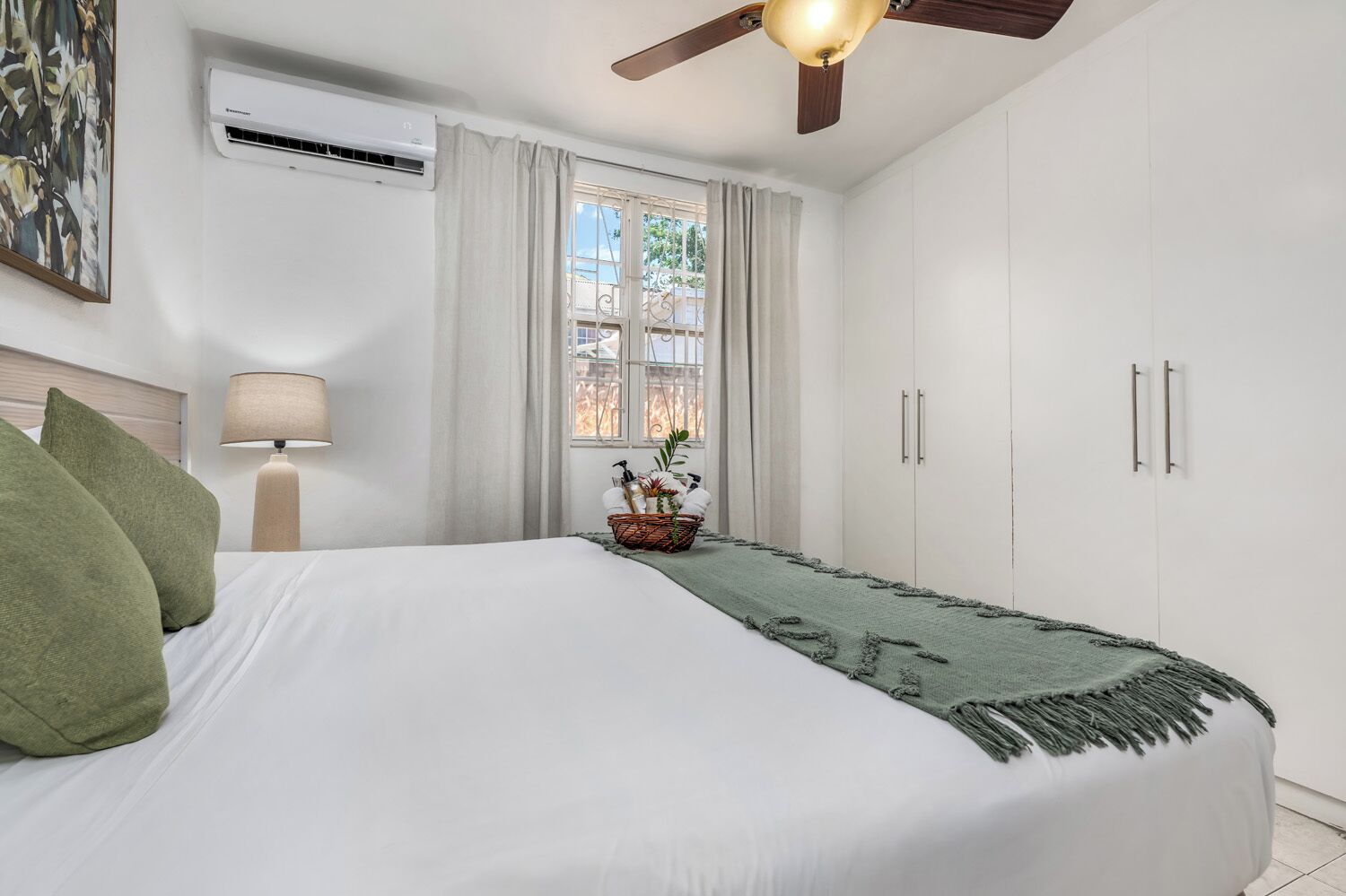 Un dormitorio con cama, ventilador de techo, ventana con cortinas, lámpara de noche y aire acondicionado instalado en la pared.
