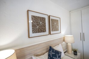 Un dormitorio con cabecera de madera, lámpara de noche, planta en maceta y dos cuadros enmarcados.