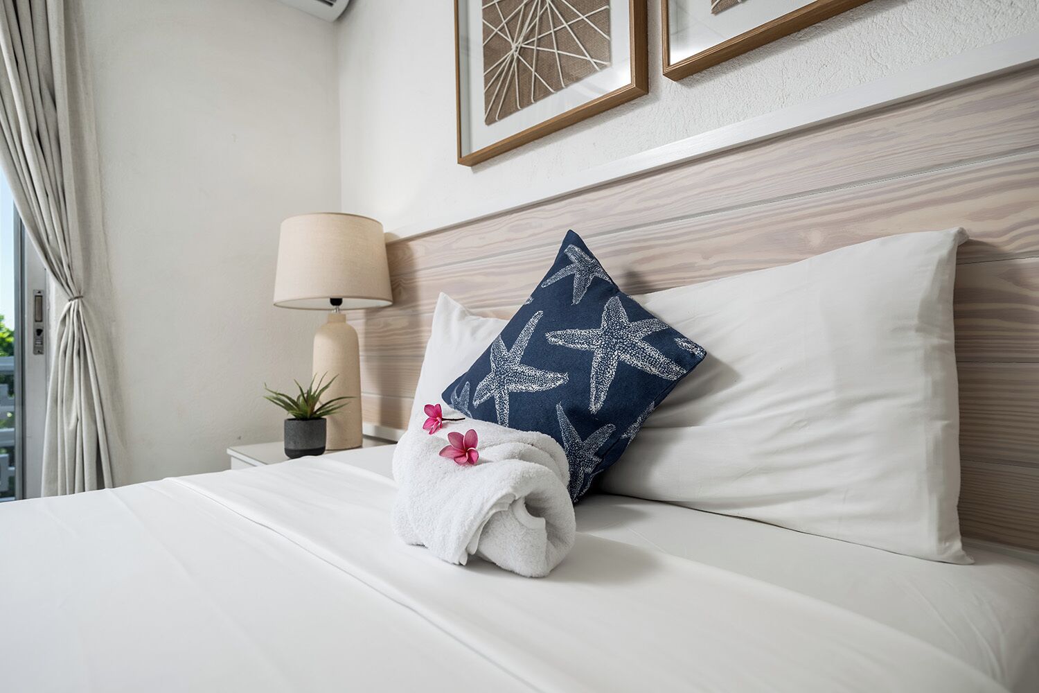 Una cama bien tendida con ropa blanca, una almohada azul con diseño de estrella de mar, una toalla blanca, una lámpara y una pequeña planta en maceta sobre la mesita de noche.