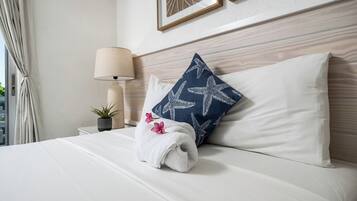 Una cama bien tendida con ropa blanca, una almohada azul con diseño de estrella de mar, una toalla blanca, una lámpara y una pequeña planta en maceta sobre la mesita de noche.