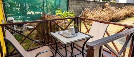 A wooden deck with a table and two chairs, a small table with a teapot and cups, and a green fence in the background.