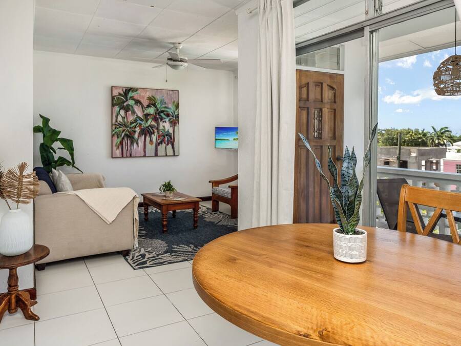 Una sala de estar con un sofá, una mesa de centro y una mesa de comedor con sillas. Hay un cuadro en la pared, una planta sobre la mesa de centro y vistas al exterior a través de una puerta.