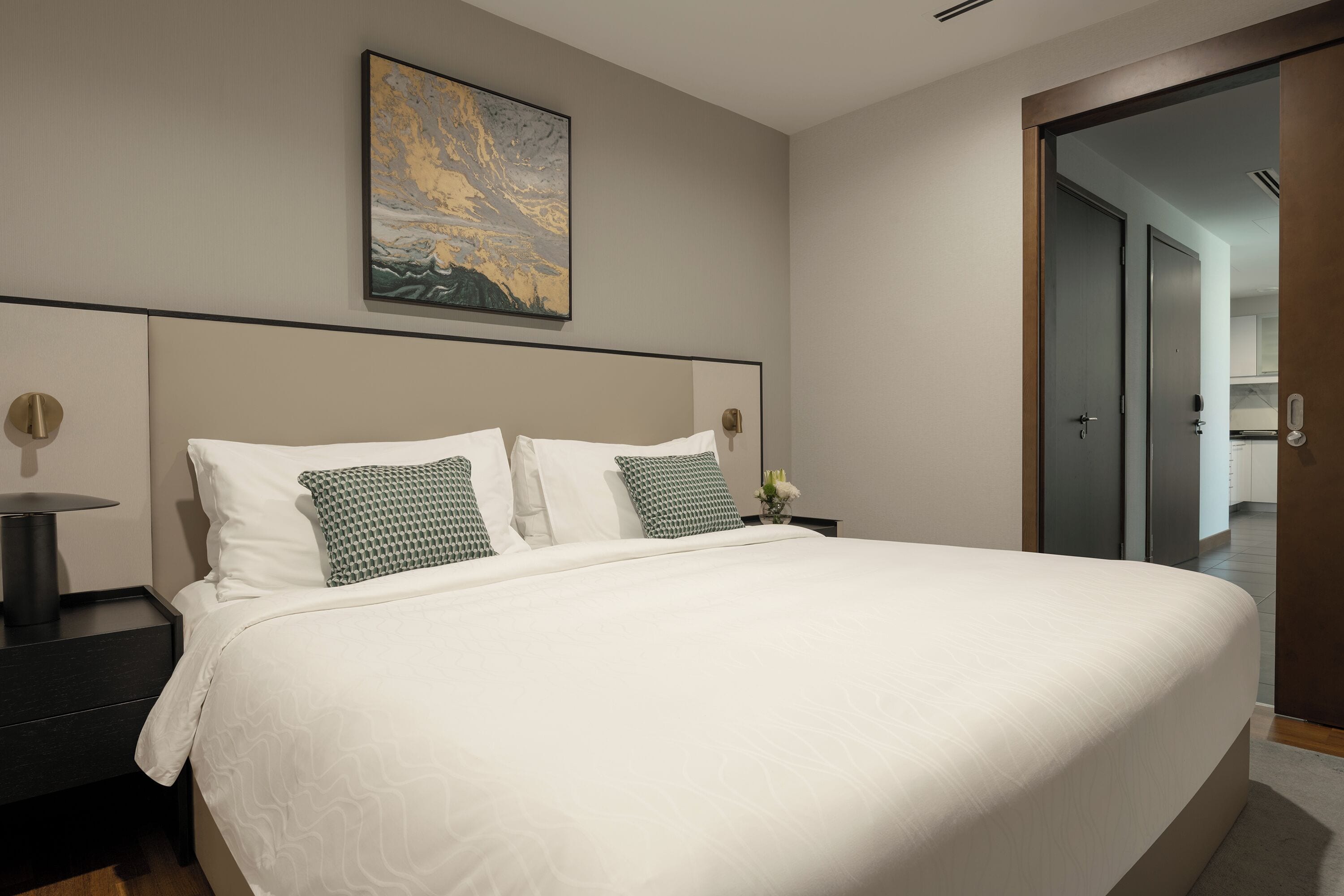 Un letto rifatto con cura, lenzuola bianche e cuscini decorati, un comodino con una lampada, un quadro appeso al muro e una porta che dà su un bagno.