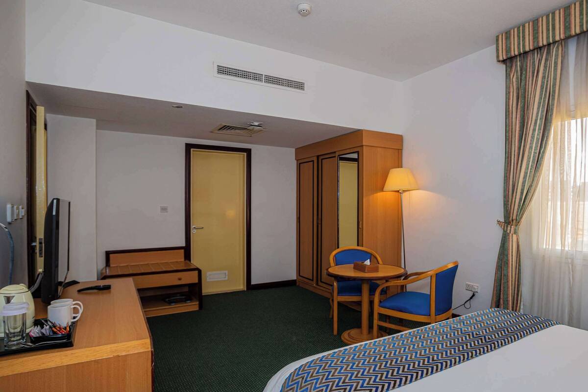 A hotel room with a bed, a desk with a computer, a chair, a table with a lamp, a TV, and a window with curtains.