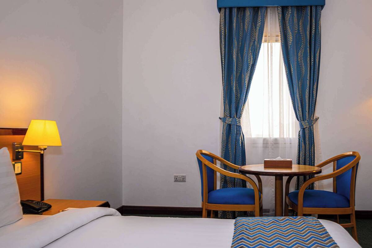 A hotel room with a bed, two chairs, a small table, a lamp, and a window with curtains.