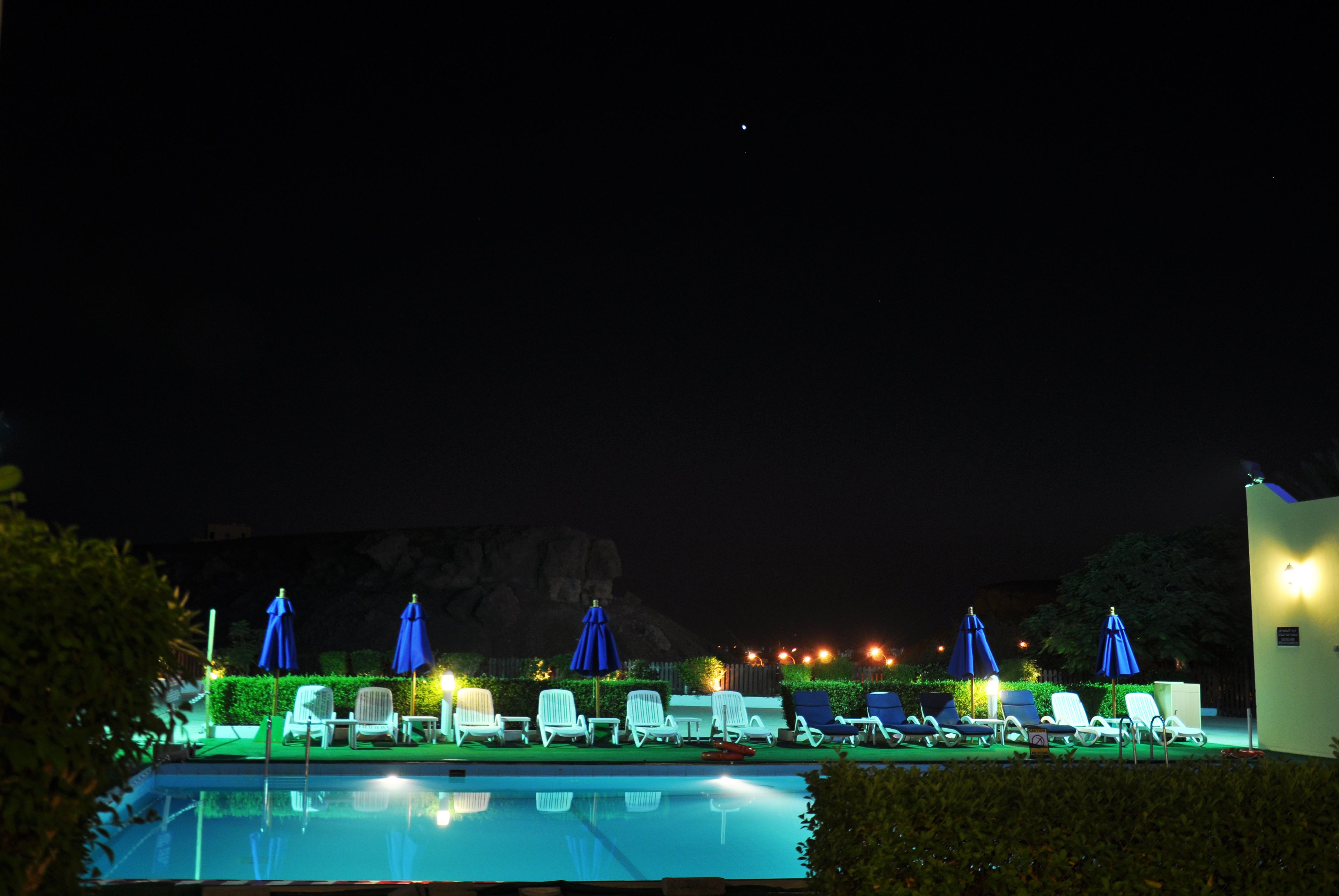 A nighttime pool area with lounge chairs and umbrellas, a clear pool, and a mountain backdrop.