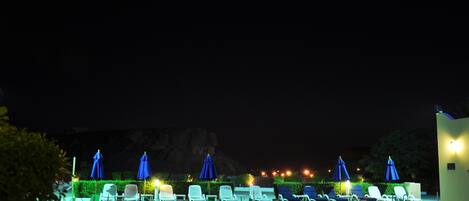 A nighttime pool area with lounge chairs and umbrellas, a clear pool, and a mountain backdrop.