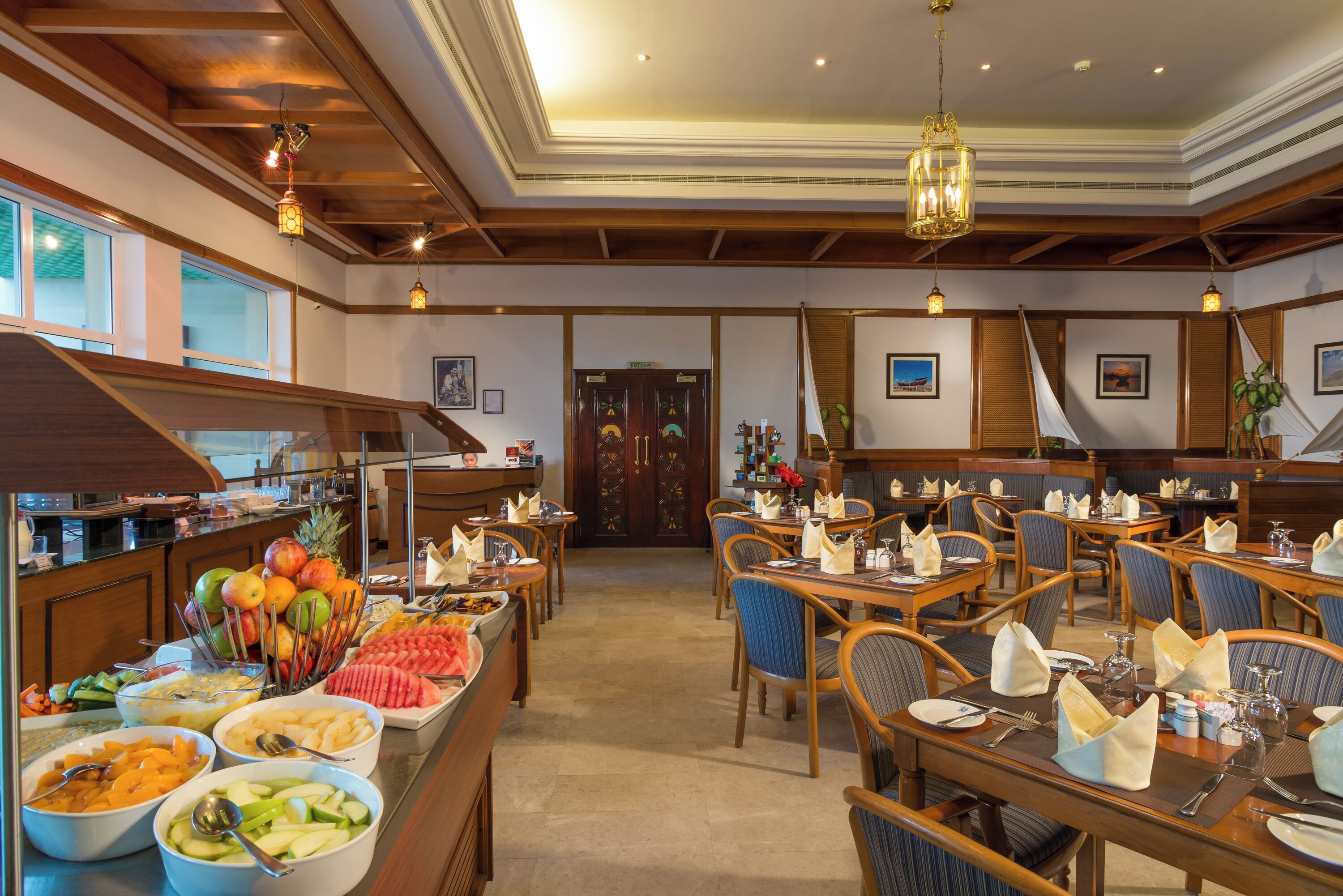 Un comedor de hotel con varias mesas dispuestas para los huéspedes, un bufé con distintos alimentos y una escalera que lleva a otro nivel.