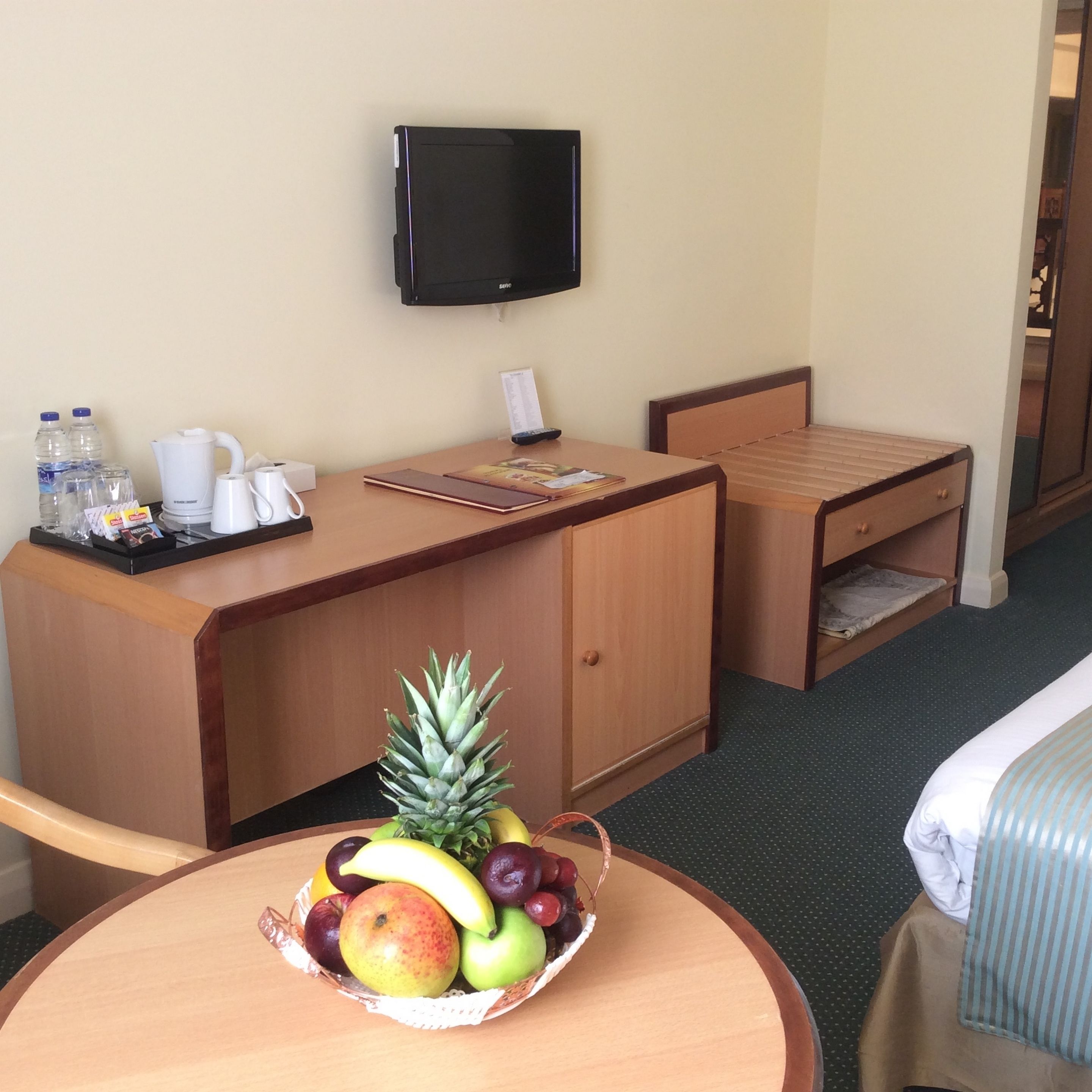 Une chambre d’hôtel équipée d’un bureau, d’un téléviseur fixé au mur, d’un lit et d’une petite table sur laquelle se trouve un panier de fruits.