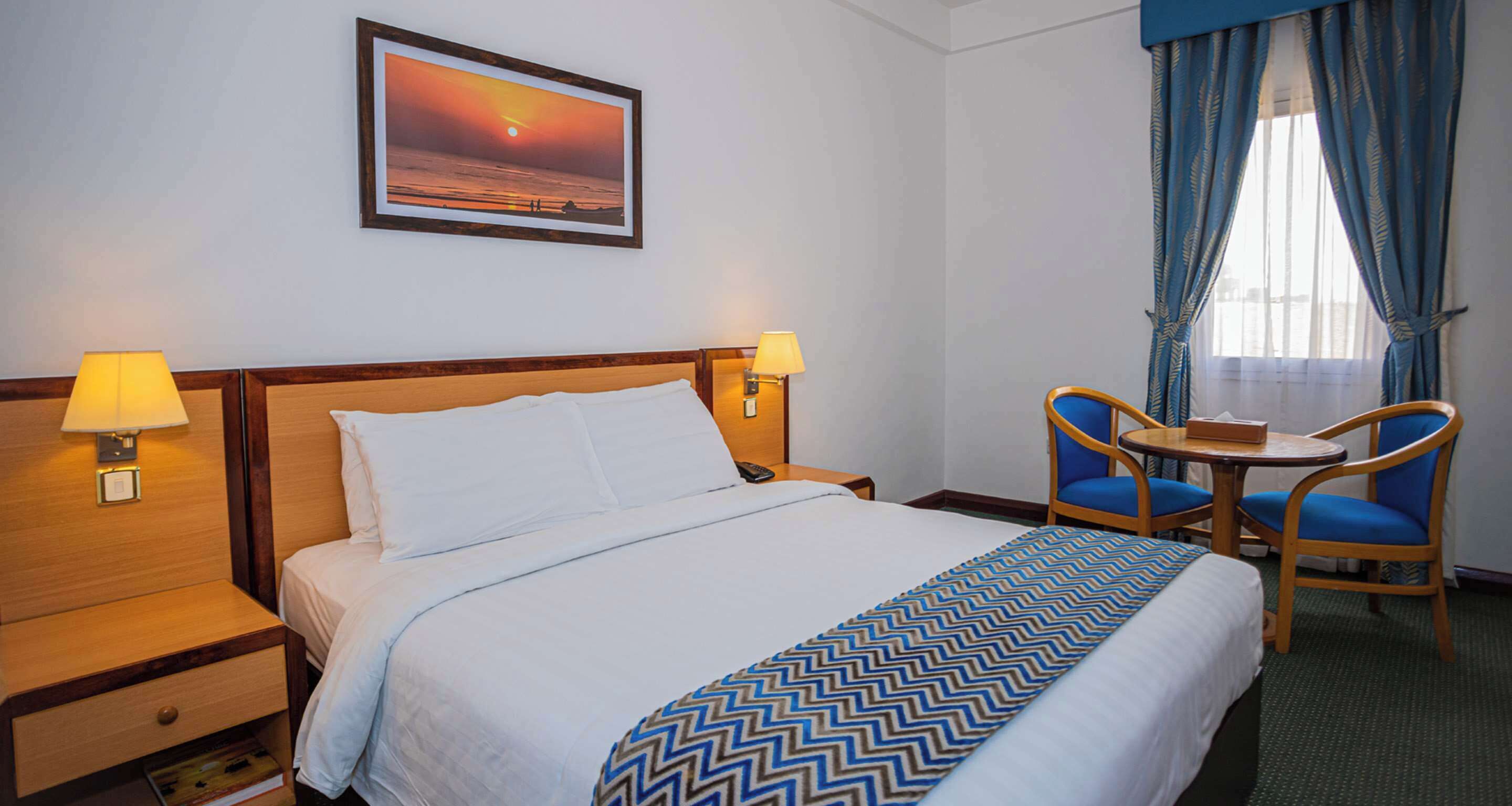 Una habitación de hotel con una cama, dos lámparas de noche, una mesa con sillas, un cuadro enmarcado en la pared y una ventana con cortinas.