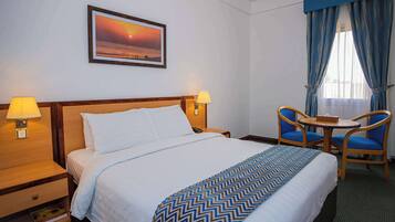 Una habitación de hotel con una cama, dos lámparas de noche, una mesa con sillas, un cuadro enmarcado en la pared y una ventana con cortinas.