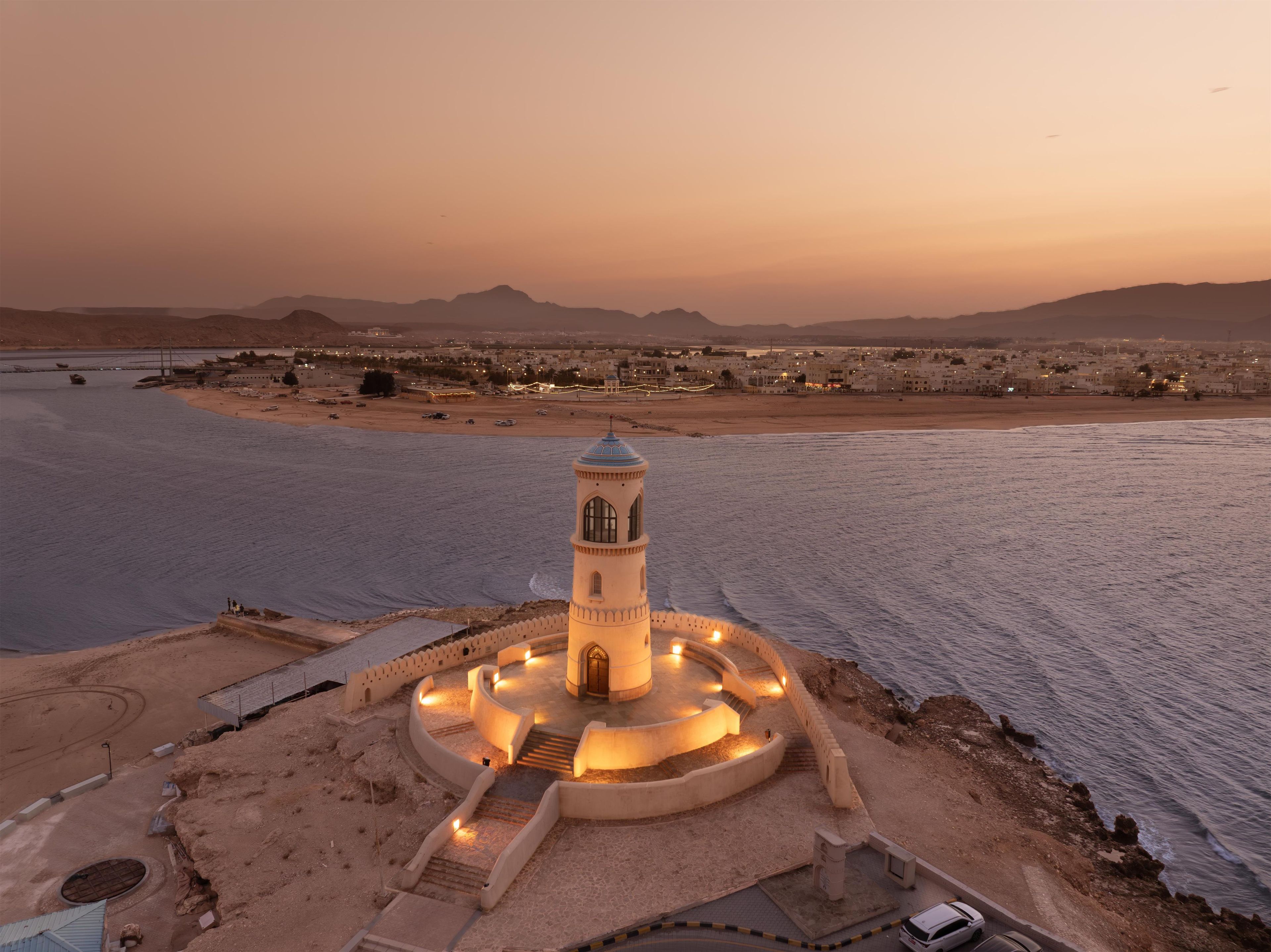 Vista costera con un faro destacado, una playa arenosa y un paisaje urbano al fondo.