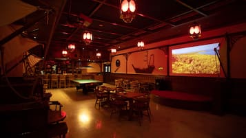 Zona de bar tenuemente iluminada con mesas de billar, sillas de madera y una pantalla grande mostrando un paisaje.