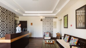 A hotel reception area with a receptionist, a reception desk, a patterned wall, a sofa with colorful cushions, and a small table with a vase and a tray. - Desert Nights Resort (Bidiya)