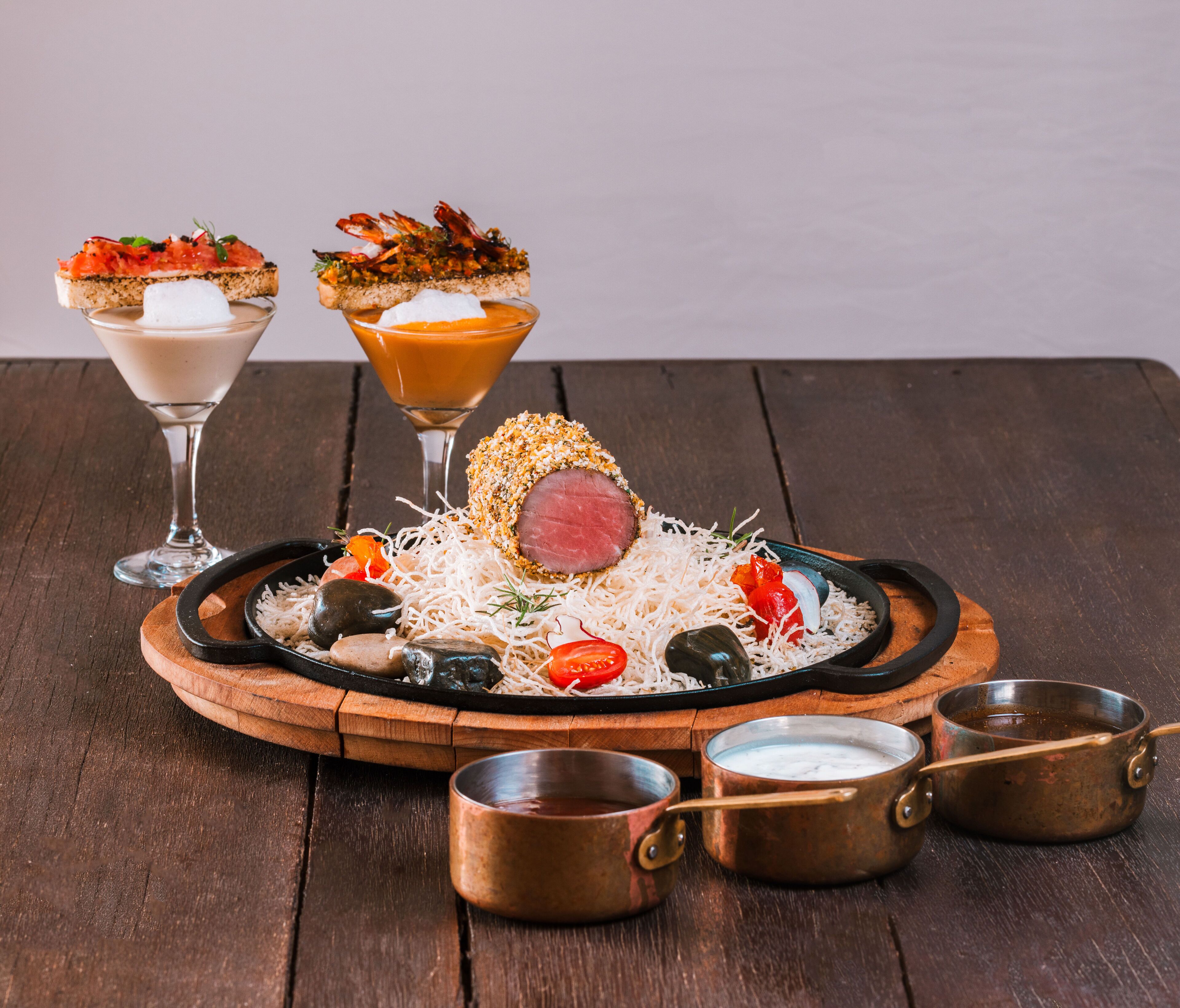 Une table en bois sur laquelle se trouvent un plat de viande, deux boissons et de petits gobelets en métal.