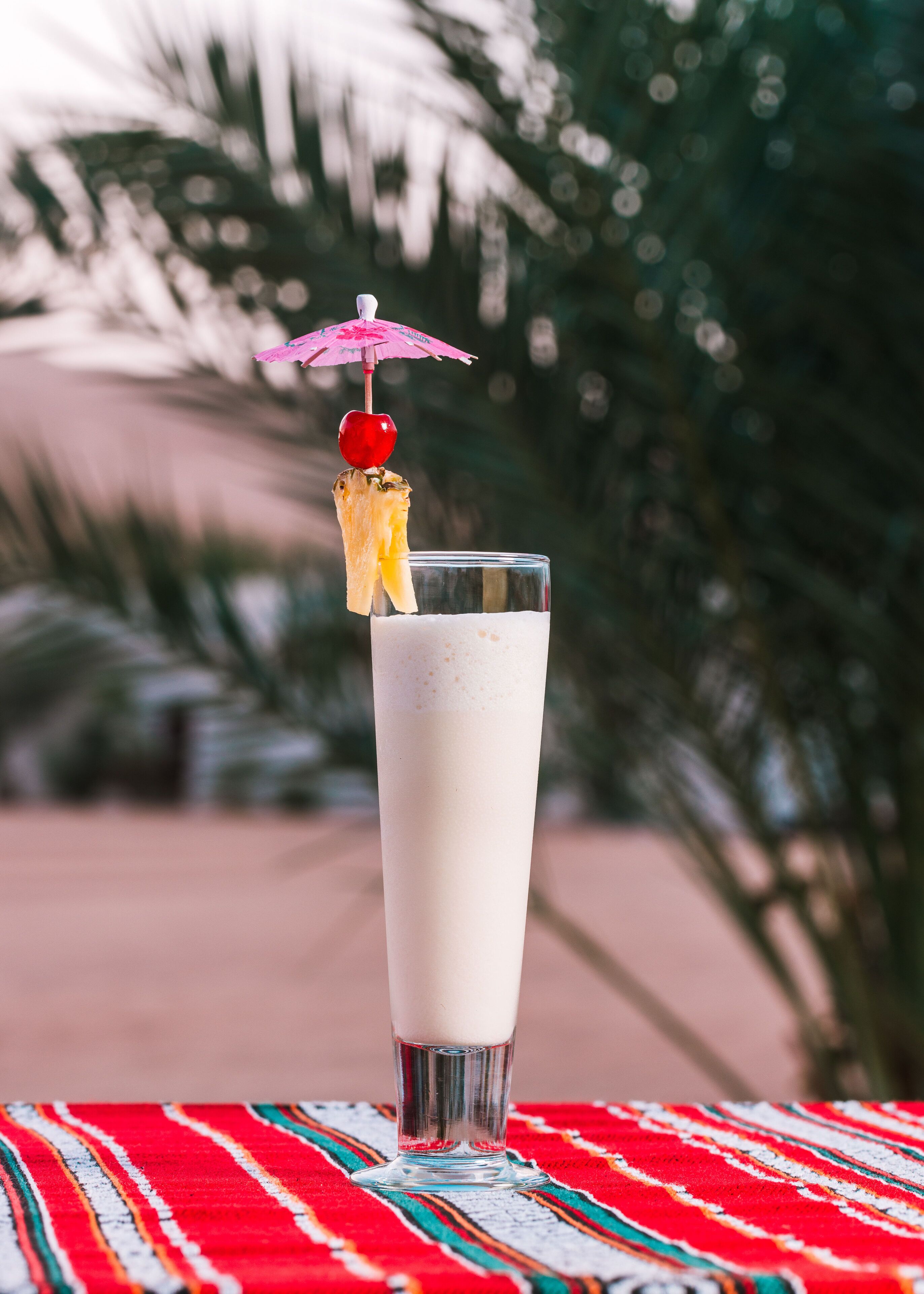 A tall glass of a creamy beverage with a red fruit garnish and a pink umbrella on a table with a red and white striped tablecloth.