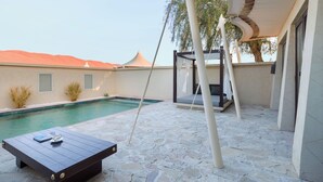 A pool area with a swing chair, a wooden table with a book and a cup, and a stone floor. - Desert Nights Resort (Bidiya)