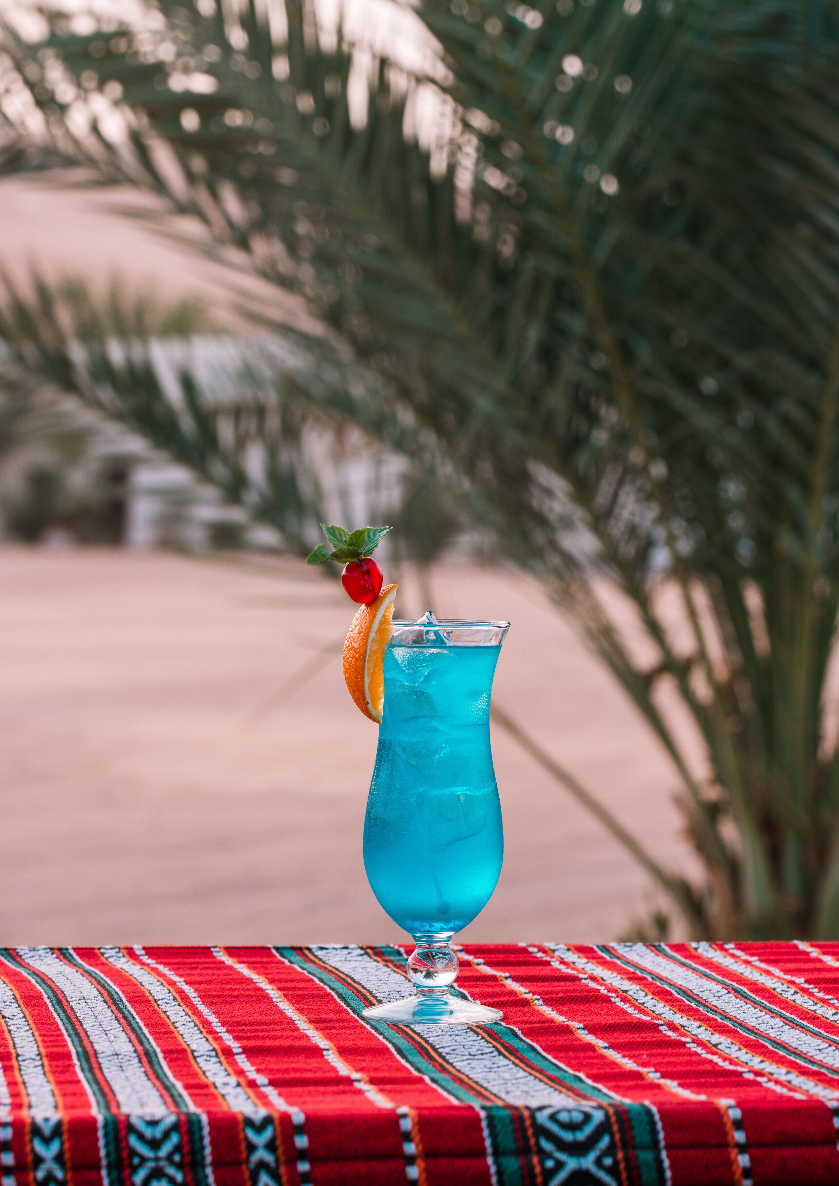 A glass of blue beverage with an orange slice and a red fruit on a red and white patterned tablecloth.