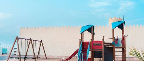 Un parque infantil con columpios y un tobogán, frente a una pared color crema y una cerca decorativa.