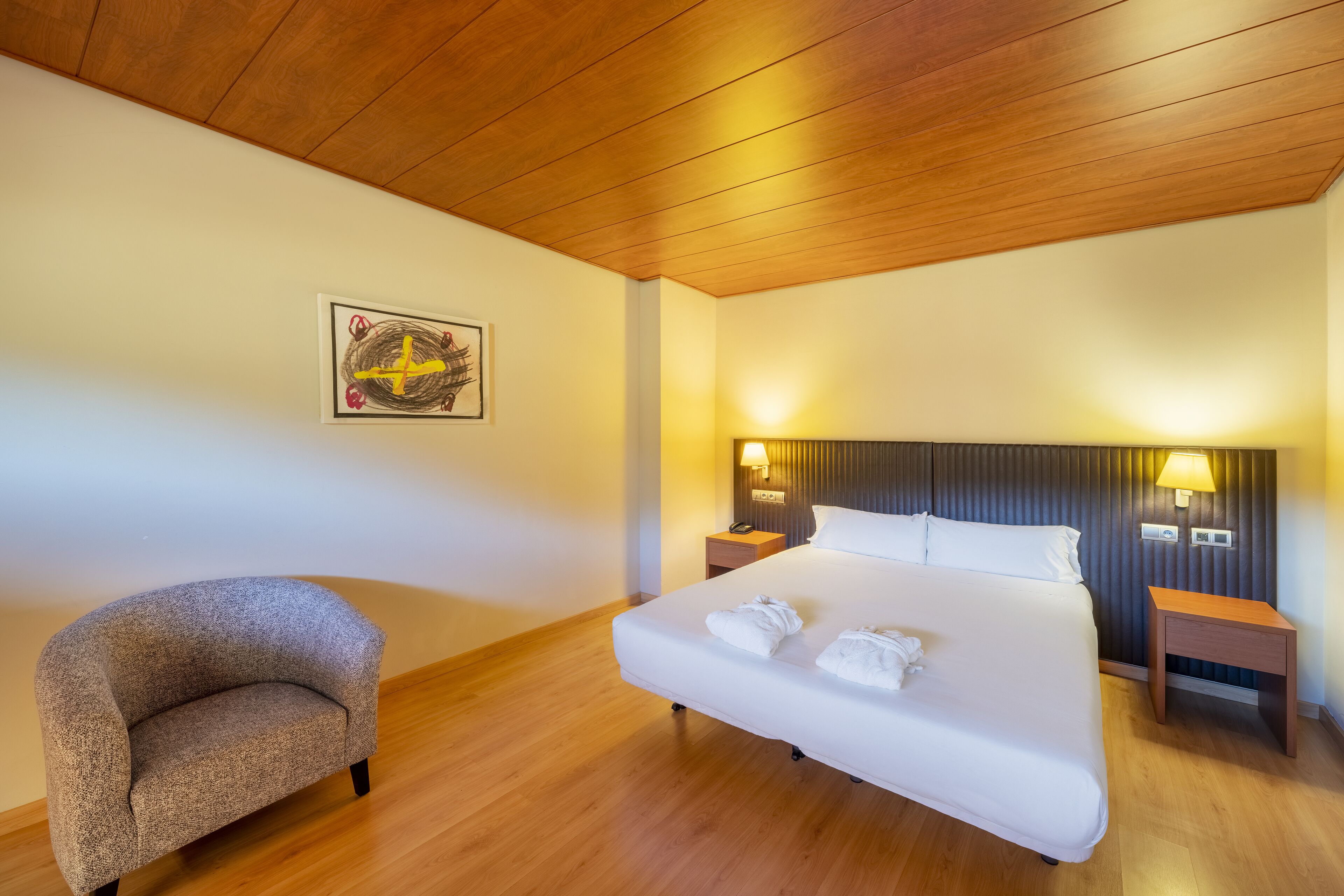 Habitación de hotel con techo de madera, una cama grande con ropa blanca, dos mesitas de noche con lámparas, un sillón pequeño y una obra de arte colgada en la pared.