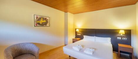 Habitación de hotel con techo de madera, una cama grande con ropa blanca, dos mesitas de noche con lámparas, un sillón pequeño y una obra de arte colgada en la pared.