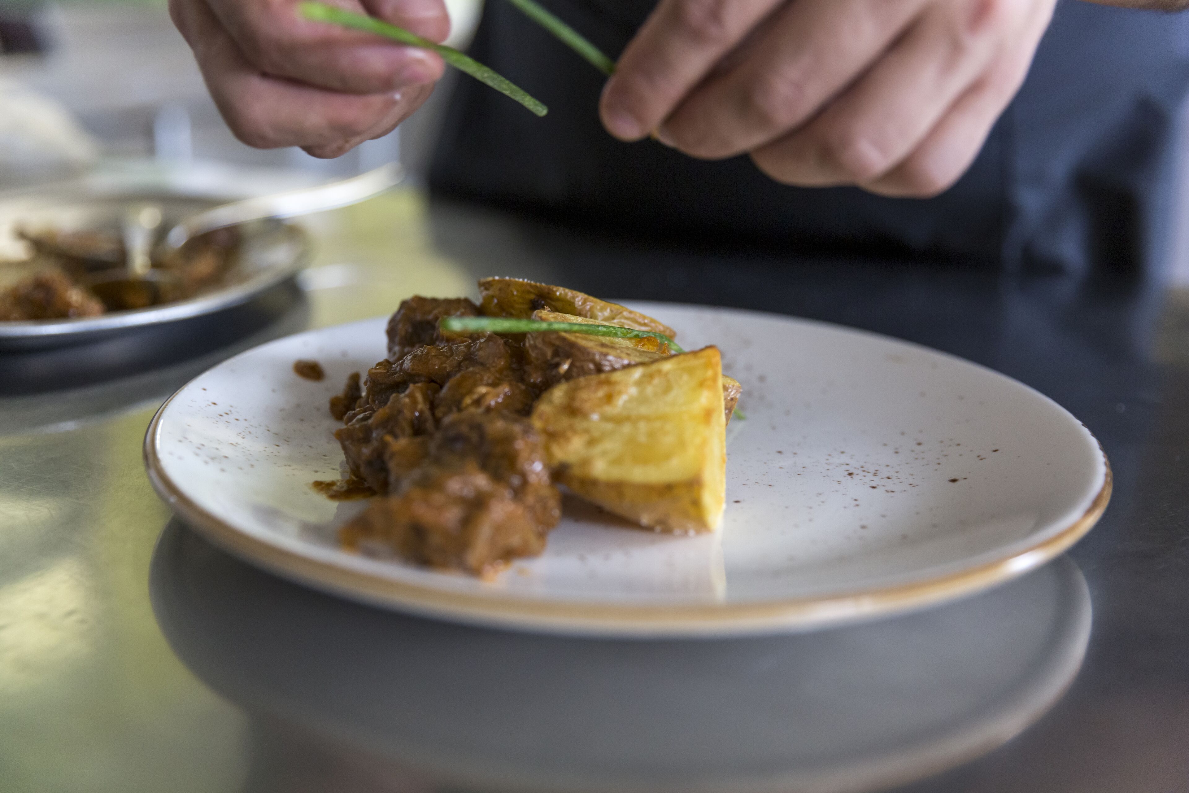 A person holding chopsticks over a plate with a piece of food and a bowl in the background.