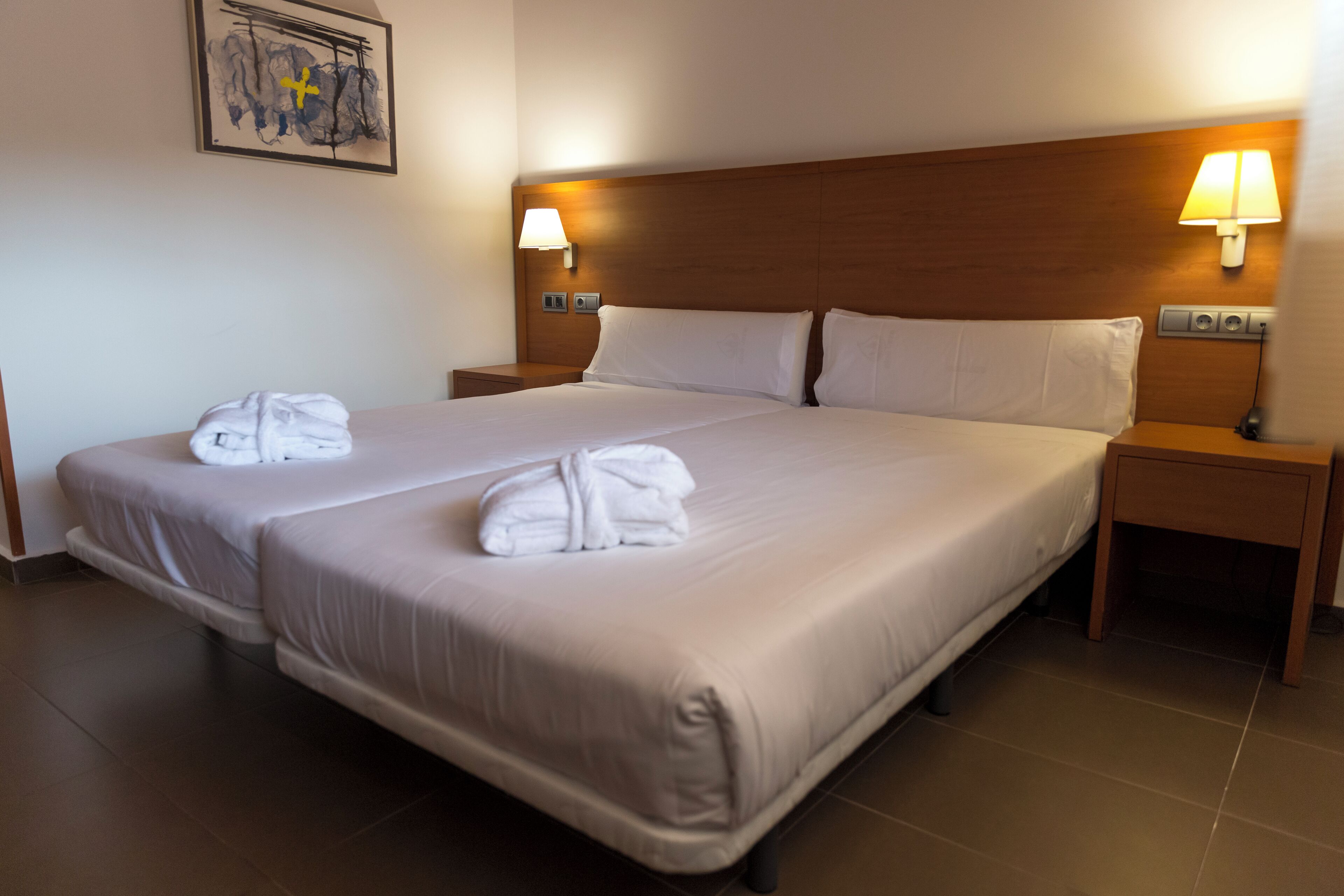 A double bed with white linens and two towels, wooden headboard, bedside lamps, and a framed picture on the wall.