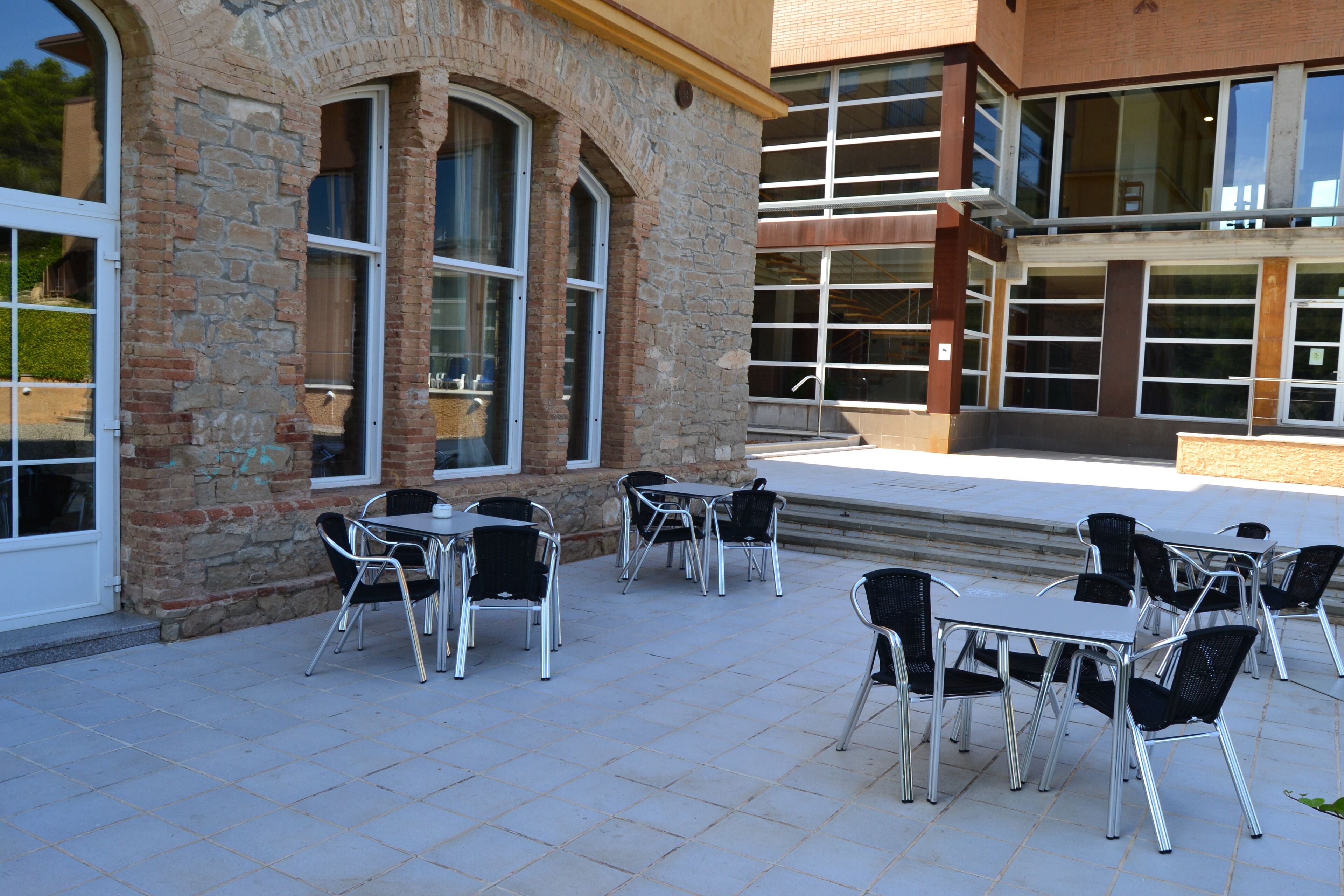 Een terras buiten met tafels en stoelen, stenen muren en grote ramen.